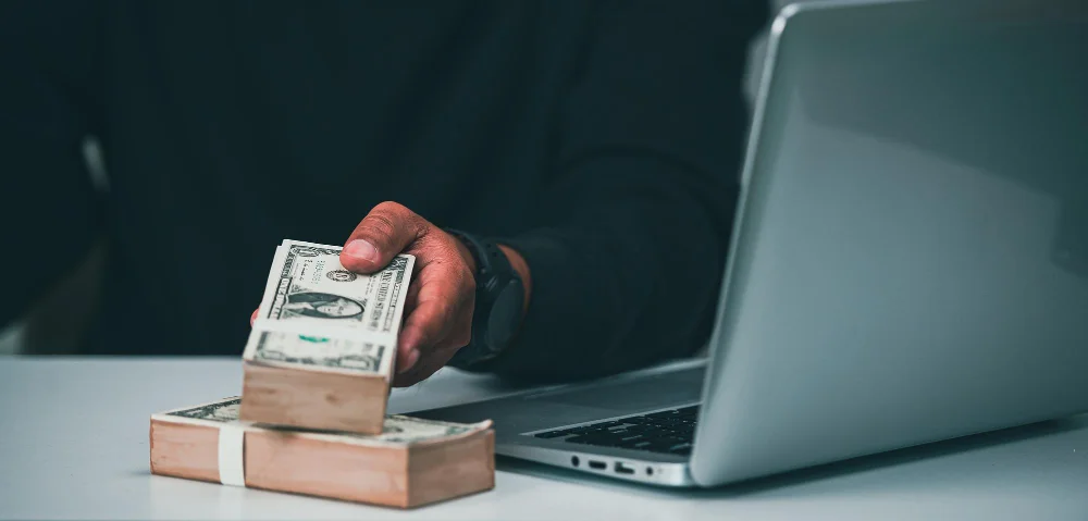 Homme tenant des liasses de billets sur un bureau avec un ordinateur portable à côté.