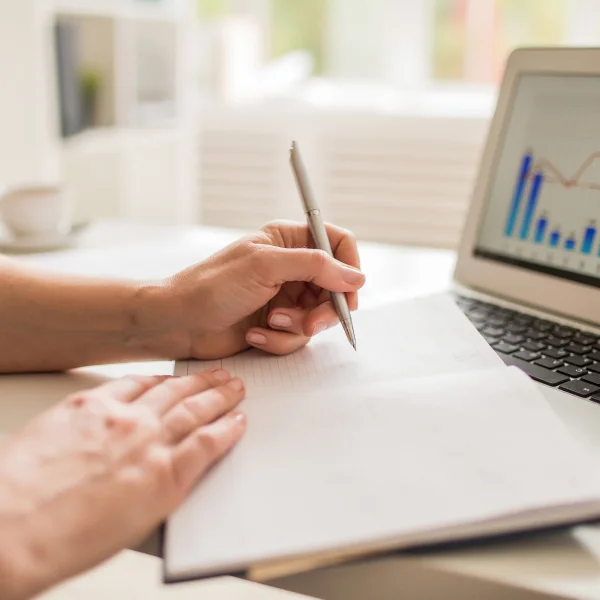 Une main de femme écrit dans un carnet avec un stylo, à côté d'un ordinateur portable, sur le thème du financement instant.