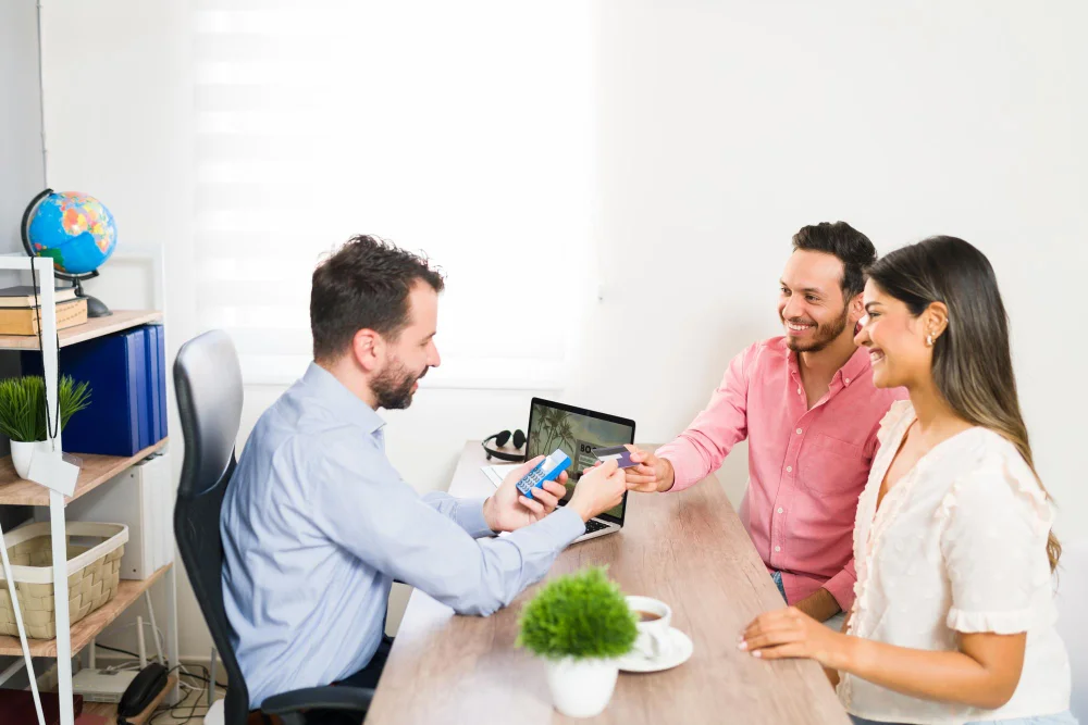 A man and woman sit at a table with a laptop; the man is focused on his phone while the woman looks at the screen.